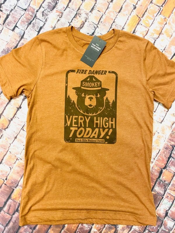 Full view of smokey's very high t-shirt design with fire danger text above smokey the bear with very high today text below and Black Hills National Forest on brick background