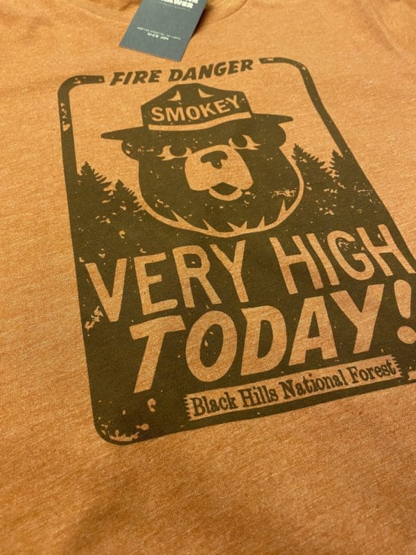 Close up of smokey's very high t-shirt design with fire danger text above smokey the bear with very high today text below and Black Hills National Forest on brick background