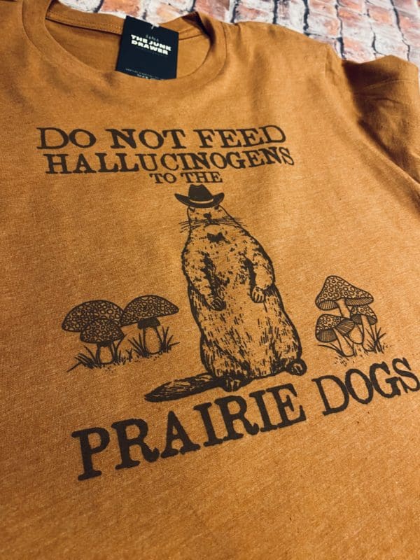 Close up view of the do not feed hallucinogens text above a prairie dog with a cowboy hat surrounded by mushrooms with prairie dogs text below t-shirt design