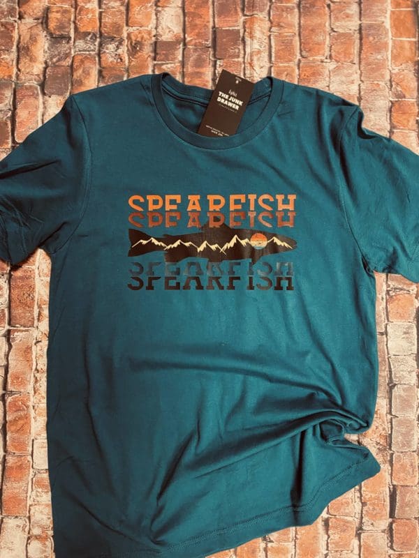 Green T-Shirt with Spearfish text with a orange to black gradient split with a fish design with mountains and a sun within