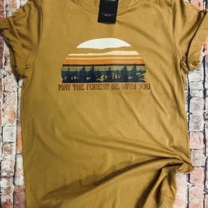 Full view of t-shirt design with a vibrant colored sun above trees with may the forest be with you text below on a light brown tee with brick background
