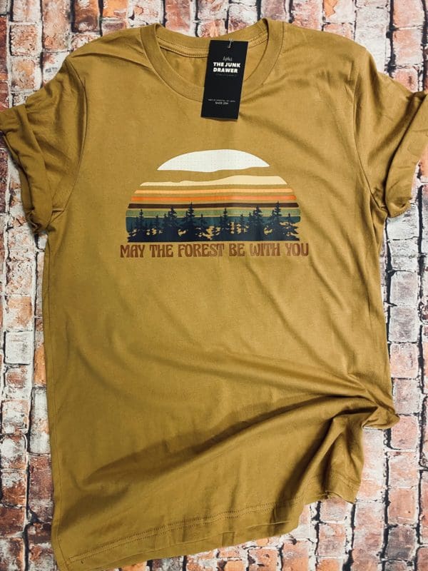 Full view of t-shirt design with a vibrant colored sun above trees with may the forest be with you text below on a light brown tee with brick background