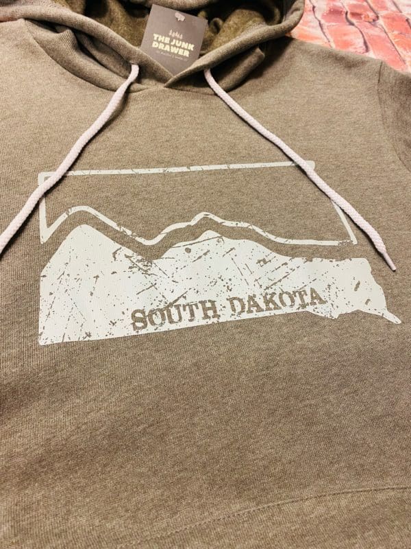 Close up view of the South Dakota state shape design split by hills in light blue on a grey heathered hoodie with contrast drawstrings