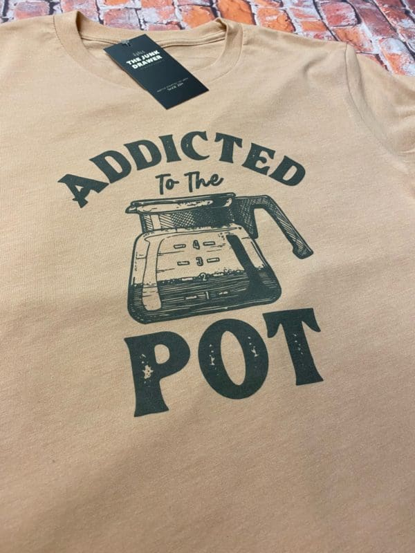 Close up of the t-shirt design with addicted to the text above a coffee pot filled less than halfway with pot below it