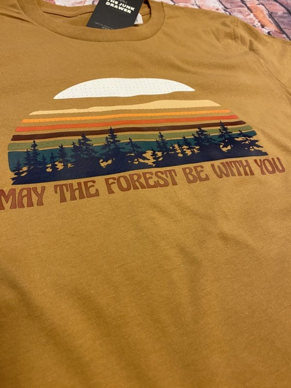 Close up view of t-shirt design with a vibrant colored sun above trees with may the forest be with you text below on a light brown tee with brick background